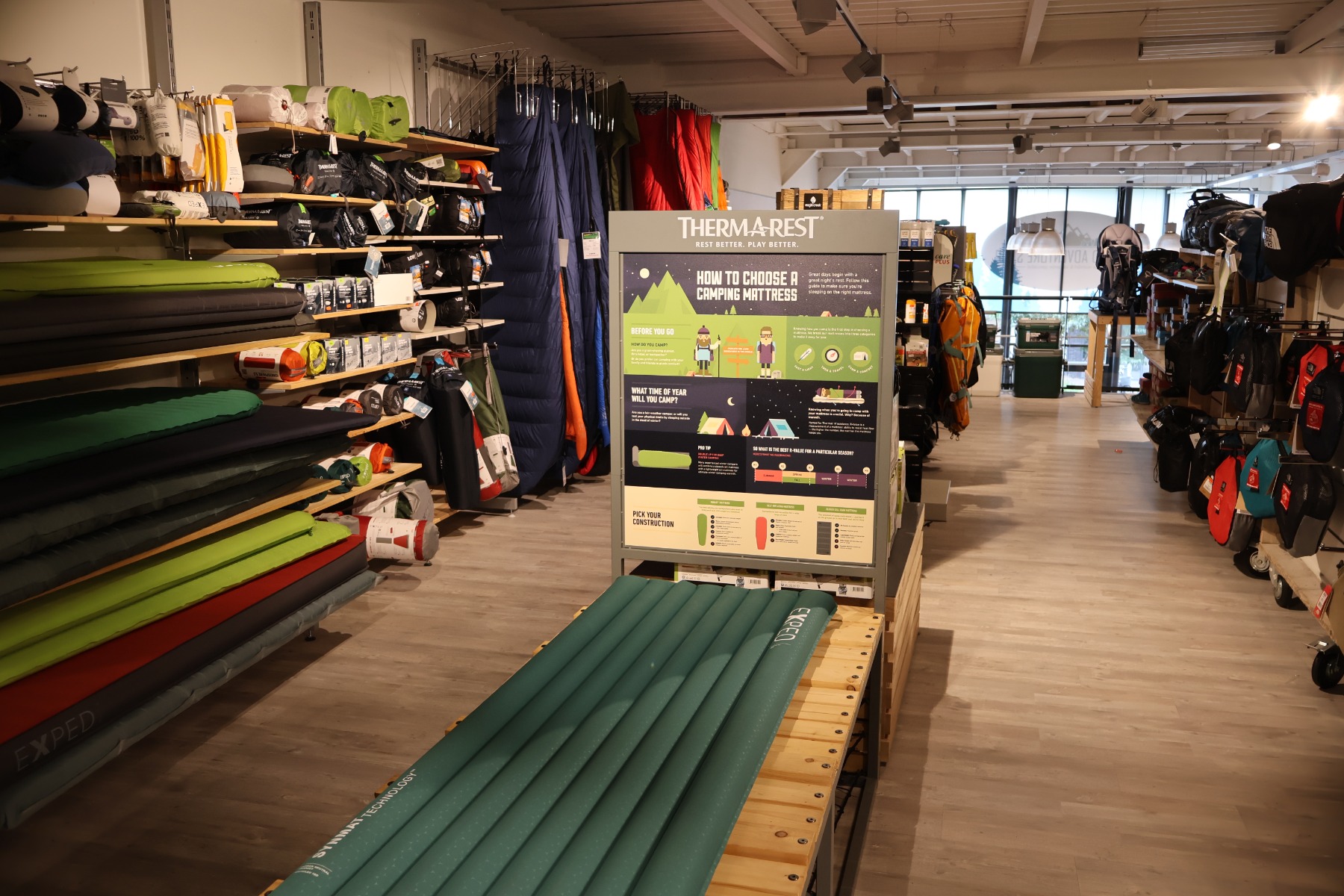 Slaapmattester in de winkel van Ronald Adventure Shop in Beverwijk, met uitlegdisplay van Therm-a-Rest en een wand vol slaapmatten en kampeeruitrusting.